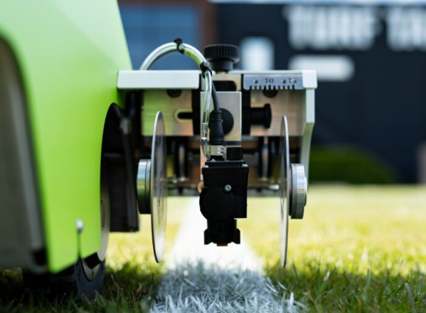 Sprüheinheit eines autonomen Markierroboters trägt präzise weiße Linienfarbe auf einen Naturrasen-Sportplatz auf.